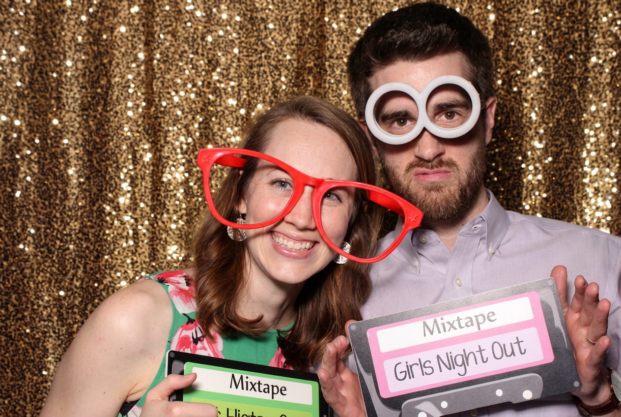 Two photo booth guests wearing oversized glasses and holding playful props in front of gold sequins